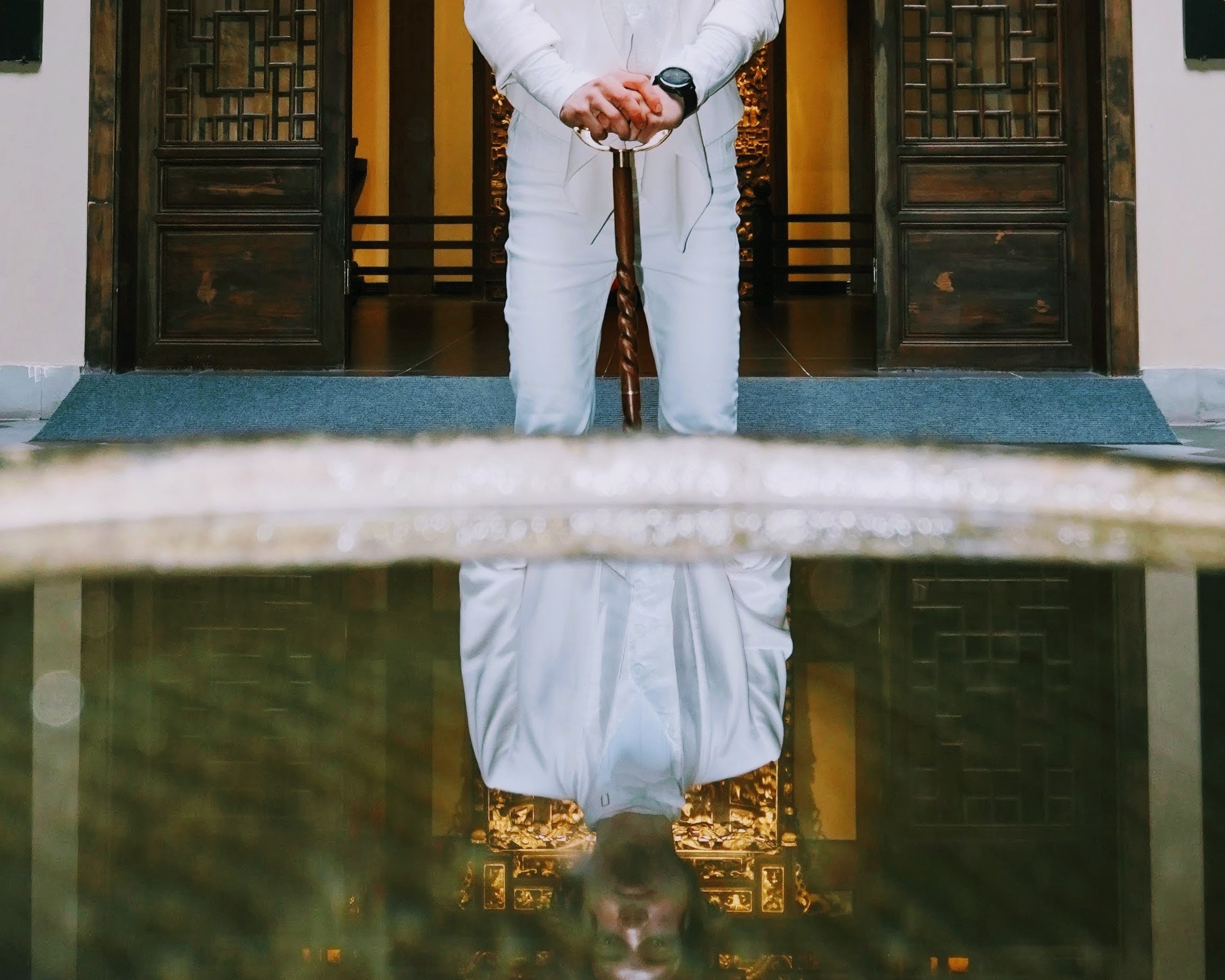 White outfit and carved wooden cane standing in front of dark wooden doors of traditional Chinese architecture, reflected in water in the foreground