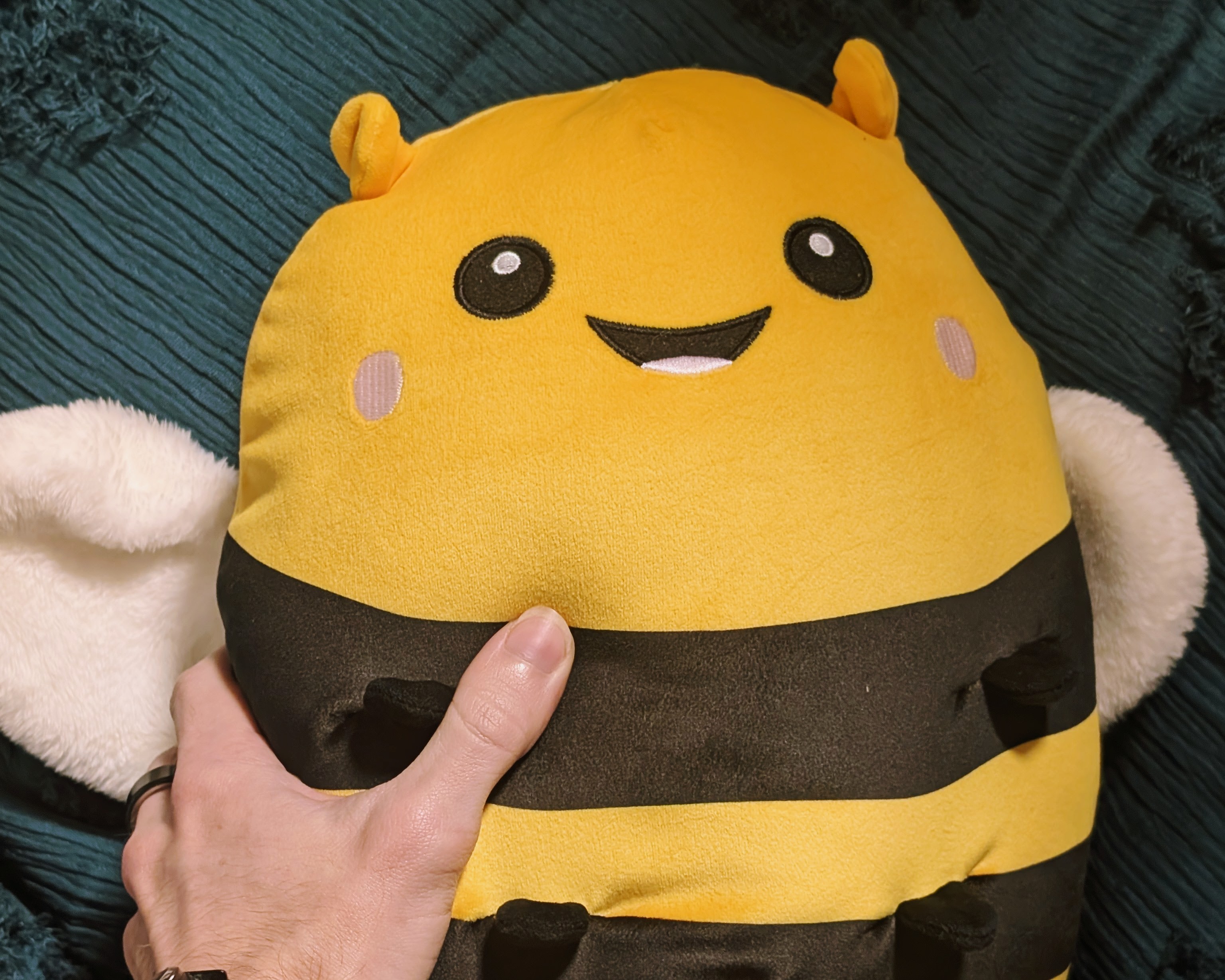 A yellow and black bee Squishmallow plushie being held against a dark green textured throw